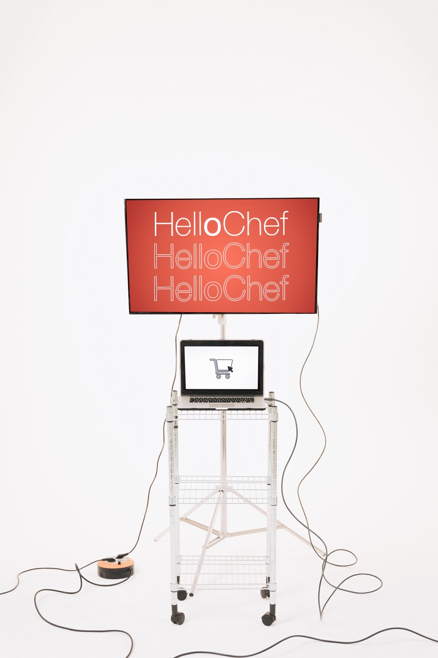 Ein Tisch mit einem Laptop, dessen Bildschirm zeigt ein Einkaufswagen icon. Dahinter ein Monitor mit rotem Hintergrund und dem HelloChef Logo.